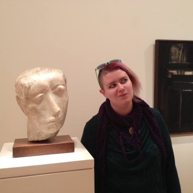 [Image Description: Ms. Fluharty is standing next to an abstract sculpted bust. She is mirroring the expression of the statue, looking to her left with her mouth skewed to the right]